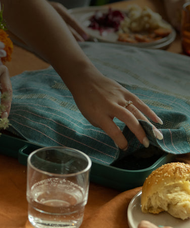 A hand with a ring lifts a blue cloth with pink stripes from a green tray on a table, with a cup of tea and a bouquet of flowers nearby.