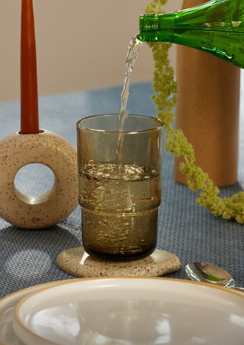 A hand pours water from a green bottle into a brown glass on a table set with plates, a spoon, a candle in a circular holder, and decorative greenery, all atop a blue tablecloth.