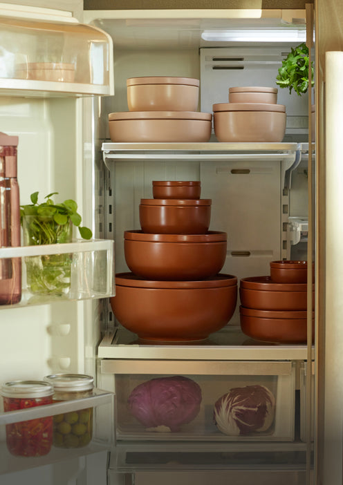 An open refrigerator filled with neatly stacked brown and beige food storage bowls, fresh herbs on the door shelf, jars of olives and peppers in a drawer, and leafy vegetables in the bottom compartment.