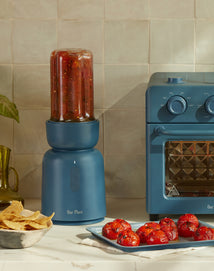 A blue blender filled with red salsa stands next to a matching blue toaster oven. In front, there’s a plate of roasted tomatoes and a bowl of tortilla chips on a white countertop.