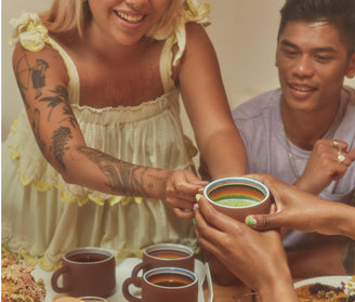 A smiling woman in a light dress with visible tattoos passes a cup to another person, while a man beside her looks on. The table is set with several cups and a platter of food.