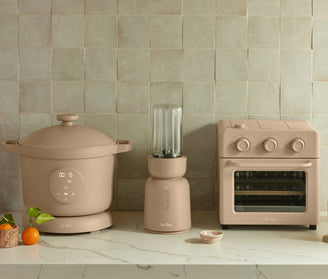 A beige slow cooker, blender, and toaster oven sit on a marble countertop against a tiled backsplash. A mandarin orange, basil leaves, and a marble bowl are also on the counter.