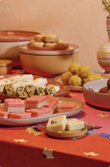 A table with assorted Indian sweets, including barfi, ladoo, and rolled sweets, displayed on plates and bowls. The tablecloth is red with decorative patterns, and a vase with flowers is in the background.