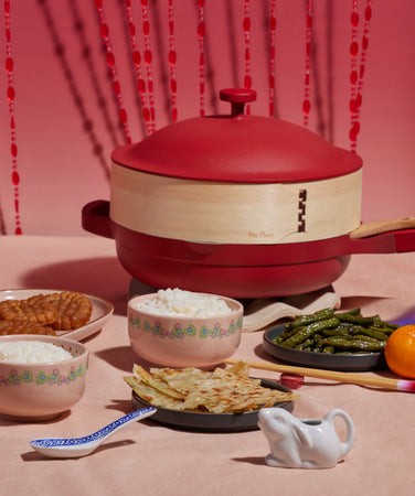A table set with red cookware, bowls of rice, plates of food, blue glasses, mandarins, candy, and flowers against a pink background with red hanging strands.