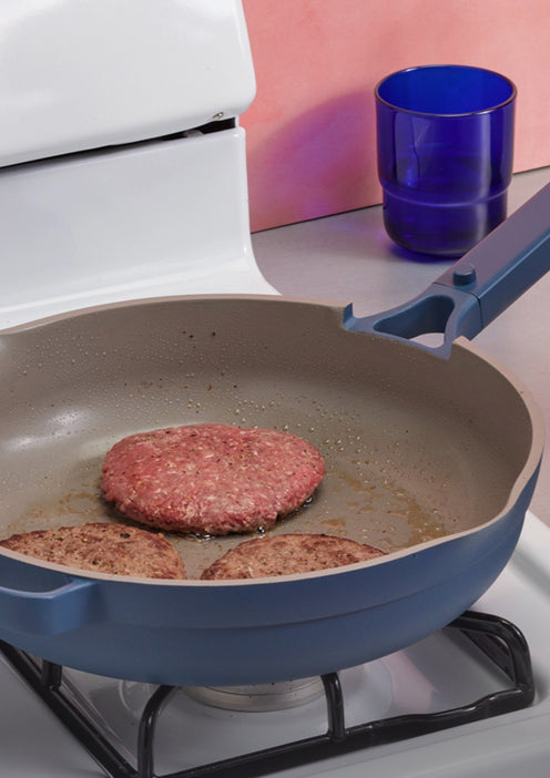 Hamburger patties cooking in a blue frying pan on a stove with a blue cup in the background.