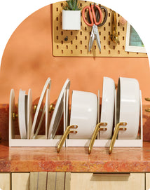 Set of white plates with gold handles in a stand on a wooden surface, with a pegboard in the background.