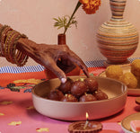 A hand adorned with red and gold bangles reaches for a gulab jamun from a bowl on a colorful table. Marigold flowers, a striped vase, and a small lit oil lamp decorate the background.