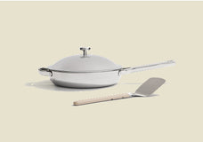 A silver frying pan with a matching domed lid and a light-colored spatula in front of it, all set against a plain beige background.