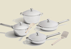 Four white pots and pans with lids are arranged on a light surface, accompanied by a white spatula and a white spoon.