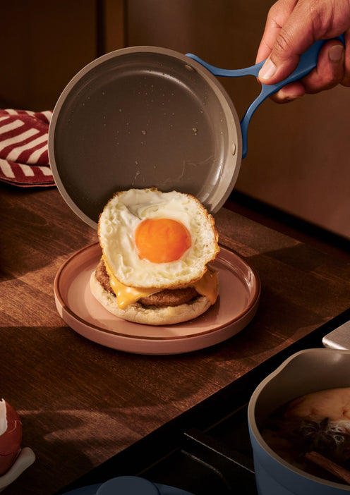 Person holding a frying pan over a burger with an egg on a wooden table.