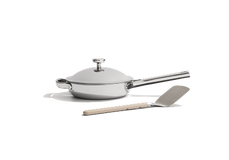 A stainless steel skillet with a lid and a metal spatula with a light-colored handle on a plain white background.