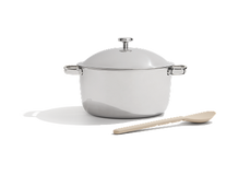 A stainless steel pot with a lid next to a light-colored wooden spoon on a white background.