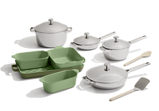 A cookware set including pots, pans with lids, green and beige baking dishes, a wooden spoon, and a spatula, arranged on a white background.