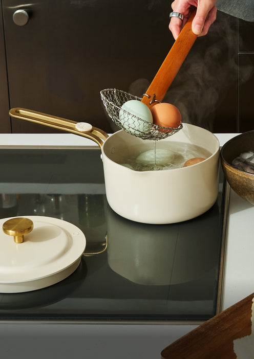 Person cooking eggs in a white pot on a stove with a wooden spoon.