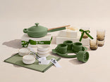 Green kitchenware set including pots, bowls, and a cutting board on a beige background.