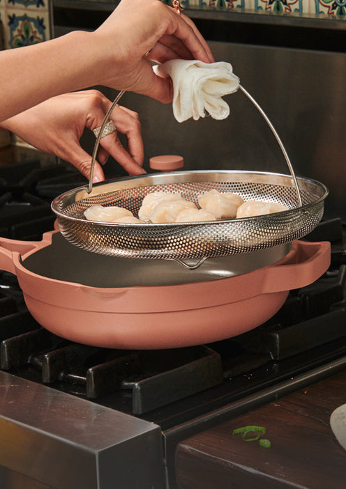 A person uses a cloth to lift a metal steamer basket filled with dumplings from a pink pan on a stovetop, with patterned tiles in the background.