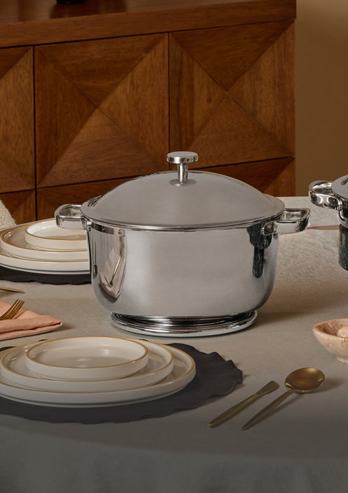 A shiny stainless steel pot with a lid is placed on a round, covered dining table set with white plates, gold utensils, and a napkin. In the background is a wooden cabinet with geometric patterns.