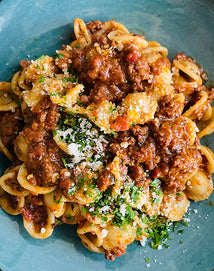 A close-up of a plate filled with pasta topped with a rich meat sauce, grated cheese, and chopped parsley, served on a blue dish.