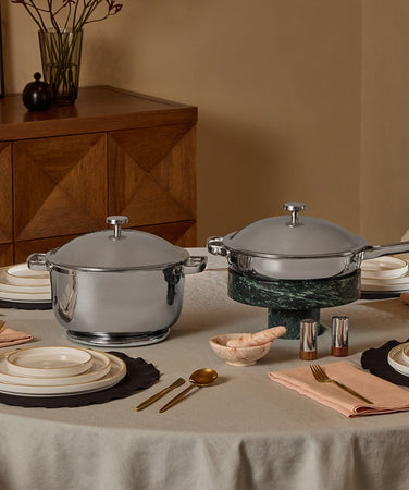 A modern kitchen countertop features a stainless steel frying pan and pot with lids, plus a marble holder containing two spatulas and a spoon. A window with greenery is visible in the background.