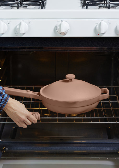 A person wearing a blue plaid shirt places a brown lidded pan on the rack inside an open oven below a white stovetop with knobs.