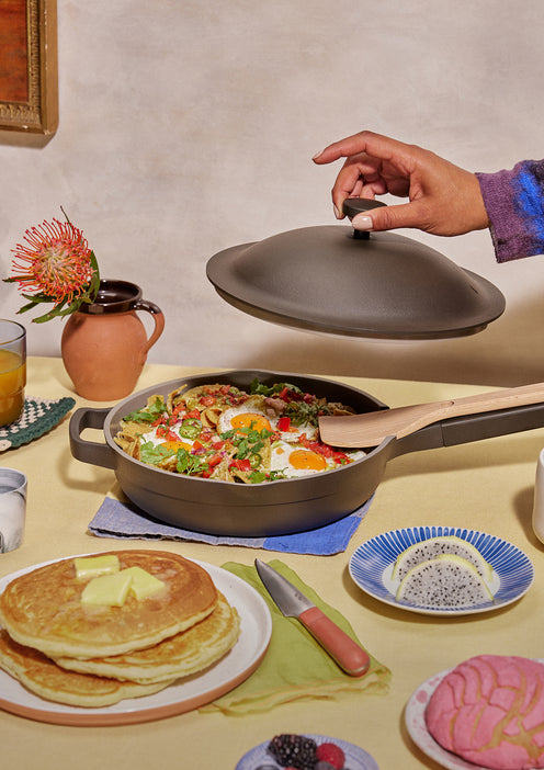 A hand lifts the lid off a skillet filled with baked eggs and vegetables. Surrounding the pan are pancakes with butter, a plate of fruit, a pink pastry, a drink, and a flower in a vase on a yellow tablecloth.
