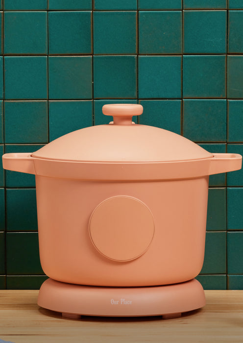 A peach-colored modern electric cooker with handles and a lid sits on a wooden countertop, against a green tiled wall background. The cooker has the brand name Our Place on the base.