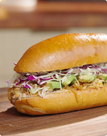 A sandwich on a shiny, golden-brown bun filled with shredded cabbage, sliced avocado, and a filling that appears to be pulled chicken, resting on a wooden surface.