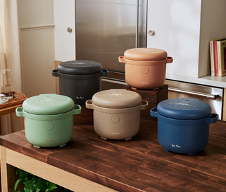 Five round electric rice cookers in different colors—black, beige, blue, green, and tan—are displayed on a wooden kitchen counter in a modern kitchen setting.