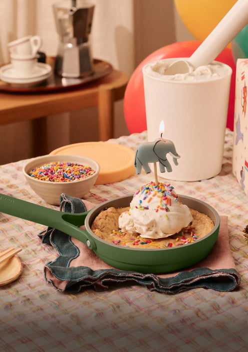 A skillet cookie topped with ice cream, sprinkles, and a lit elephant-shaped candle sits on a table beside a bowl of sprinkles, a tub of ice cream, and a coffee maker in the background.