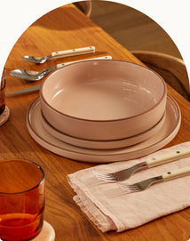 A set of pink ceramic dishes, including a bowl and two plates, is neatly arranged on a wooden table with stainless steel cutlery on a pale napkin and amber glassware nearby.