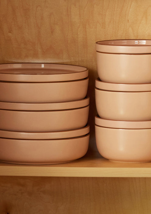 Stacked tan ceramic plates and bowls with brown rims are arranged neatly on a wooden shelf.