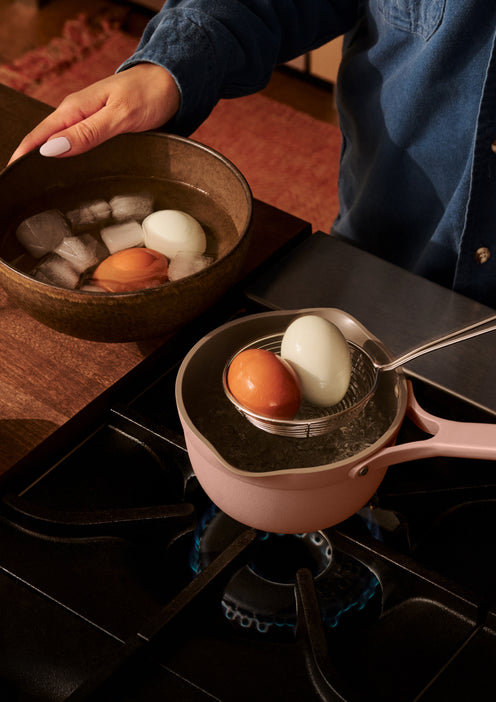 A person transfers boiled eggs from a saucepan on a stove to a bowl of ice water using a slotted spoon.