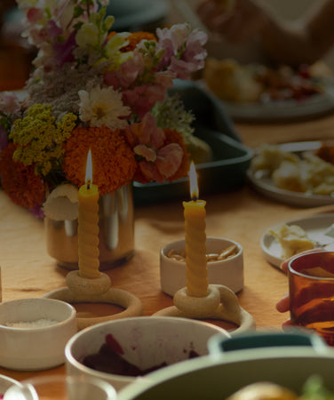 A table set for a meal with plates of food, a hand holding a glass, two lit candles in holders, and a vase of colorful flowers as a centerpiece.