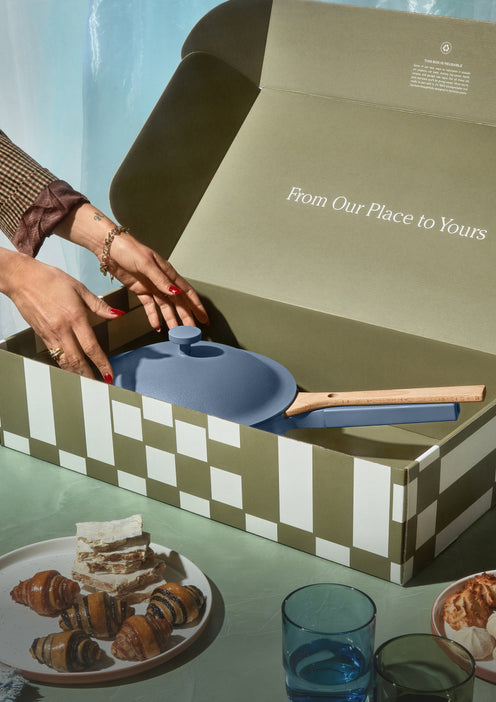 A person opens a large checkered box displaying a blue pan with a wooden handle. The inside of the box reads “From Our Place to Yours.” Nearby, a table holds pastries, desserts, and glass cups.