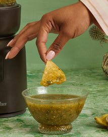 A hand dips a tortilla chip into a glass bowl of green salsa, with a blender visible on the left and a green marbled surface and background.