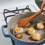 A person uses a wooden spatula to sear scallops in a blue skillet on a gas stove; the scallops are browning on one side and there are small puddles of oil in the pan.