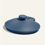 A blue, round, silicone hot water bottle with a screw cap, lying flat against a light beige background.