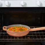 A tan frying pan with a handle sits on an oven rack, baking an egg dish garnished with green vegetables, in front of a white stove control panel.