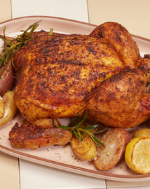 A whole roasted chicken with crispy, seasoned skin is served on a platter with roasted potatoes, yellow vegetables, and sprigs of rosemary.