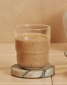 A glass of thick, light brown smoothie with visible specks, placed on a round, marbled coaster against a beige background.