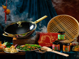 A steaming black and gold wok sits next to a bamboo steamer, jars of sauces, a red box with dragon design, wooden utensils, plates of dumplings and greens, and teacups, all arranged on a velvet blue cloth.