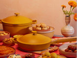 A table set with yellow cookware, various traditional sweets and snacks, and a striped vase with orange marigold flowers, against a peach-colored textured wall.