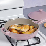 A pink nonstick skillet with a matching lid beside it is on a gas stove, cooking seasoned chicken drumsticks, lemon slices, and fresh herbs.