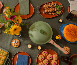 A dinner table set with a green pot, cornbread in a blue dish, skewered grilled meat, floral arrangement, cupcakes, drinks, and plates on a green tablecloth. Candles are scattered around the table.