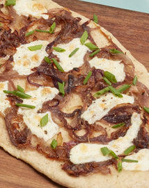 A close-up of a flatbread topped with caramelized onions, melted cheese, and chopped green onions on a wooden serving board.