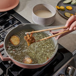 A hand uses chopsticks to fry battered vegetables in hot oil in a pink pan on a stove. Sliced vegetables and a bowl of batter are nearby on the counter.