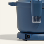 A close-up of a blue electric kitchen appliance, likely a rice cooker or pressure cooker, showing part of the lid, handle, power port, and a clear measuring cup attached to the side.