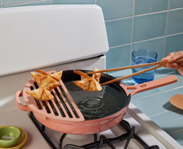 A hand uses chopsticks to fry and lift crispy wontons from a pink skillet filled with oil on a stove; a cooling rack rests on the pan, and a glass of water sits nearby.