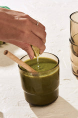 A hand squeezes a slice of lime over a glass of green smoothie with a wooden spoon inside. Two stacked brown-tinted glasses are on the right against a light textured surface.
