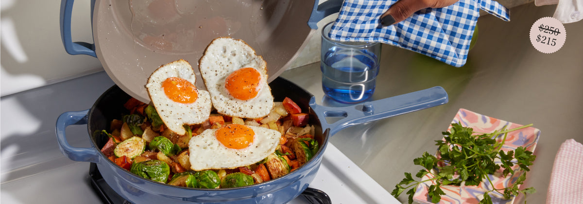 Translation missing: en.A hand adds three sunny-side-up eggs from a pan onto a skillet filled with vegetables like Brussels sprouts and potatoes on a stovetop. A blue checkered oven mitt, fresh herbs, and glass of water are nearby